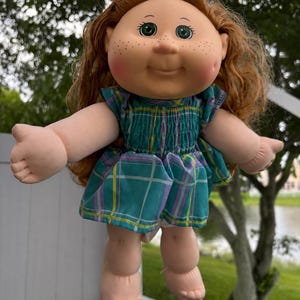 May include: A doll with long wavy auburn hair, a teal hair bow, and green eyes. The doll has a round face with freckles and rosy cheeks. It wears a sleeveless teal, purple, and yellow plaid dress with a smocked bodice and ruffled shoulders.