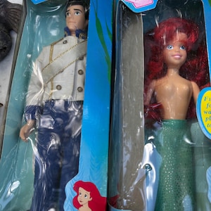 May include: Two Disney dolls in their original packaging. One doll is Prince Eric, wearing a white jacket with gold trim and blue pants. The other doll is Ariel, with red hair and a green mermaid tail. Both boxes have the Disney's logo.