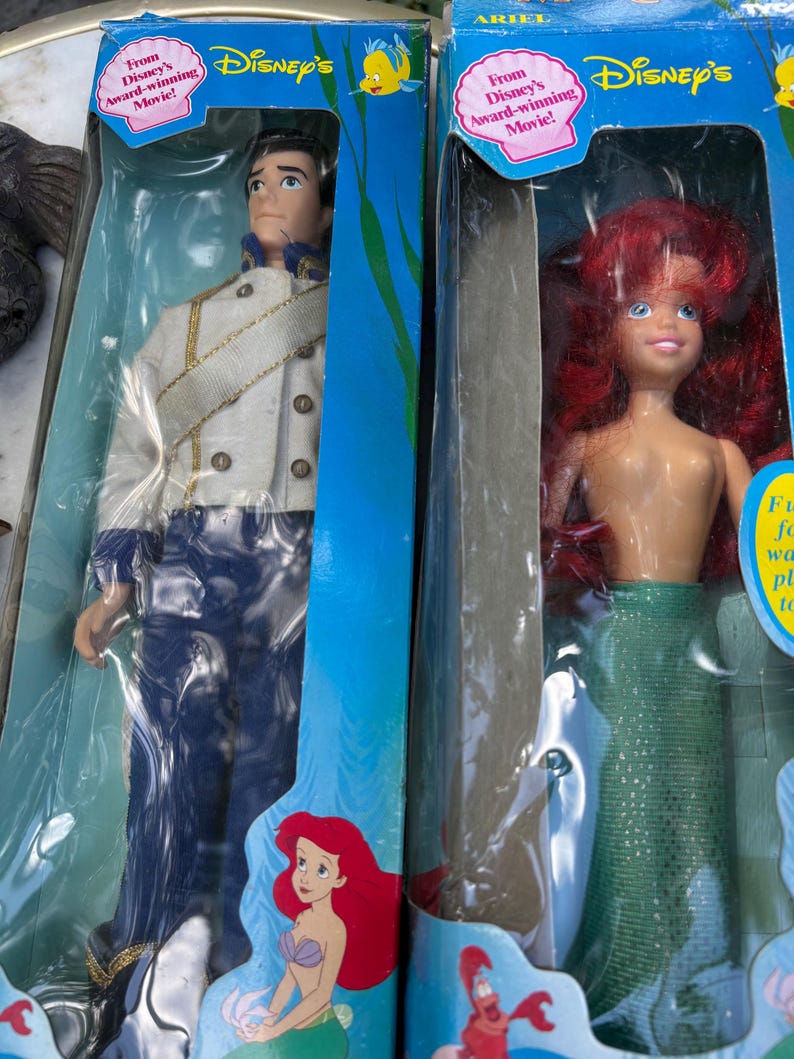 May include: Two Disney dolls in their original packaging. One doll is Prince Eric, wearing a white jacket with gold trim and blue pants. The other doll is Ariel, with red hair and a green mermaid tail. Both boxes have the Disney's logo.