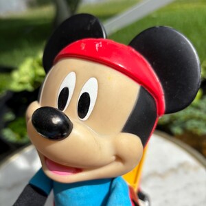 May include: A Mickey Mouse figurine wearing a red headband, blue shirt, and black gloves. The classic cartoon character has black ears, a black nose, and white eyes. The figurine is a collectible item.
