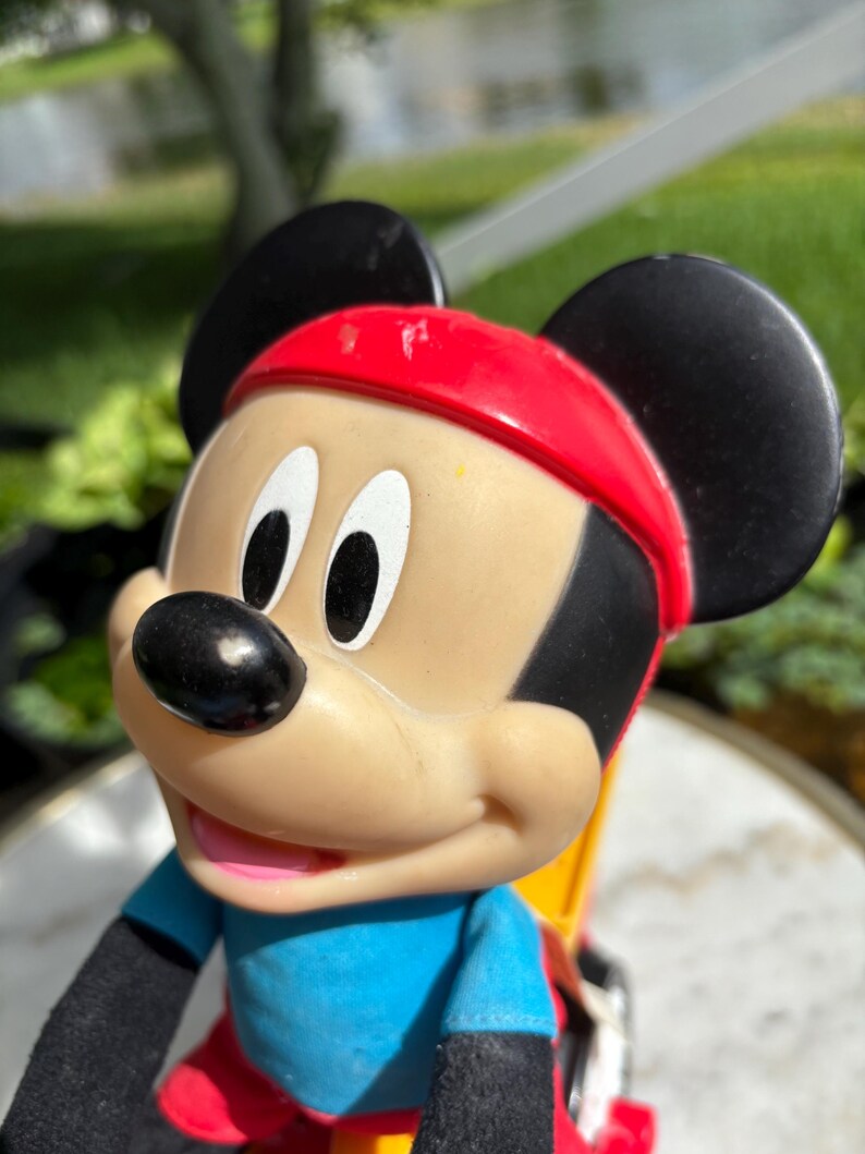 May include: A Mickey Mouse figurine wearing a red headband, blue shirt, and black gloves. The classic cartoon character has black ears, a black nose, and white eyes. The figurine is a collectible item.