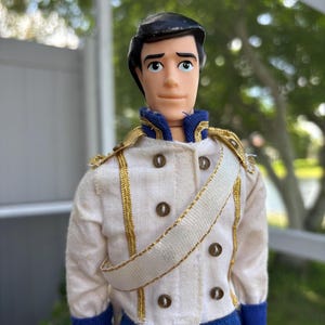 May include: A vintage doll of a prince, wearing a white jacket with gold trim and dark brown buttons. The jacket has a white sash and blue accents. The doll has black hair and is looking forward.