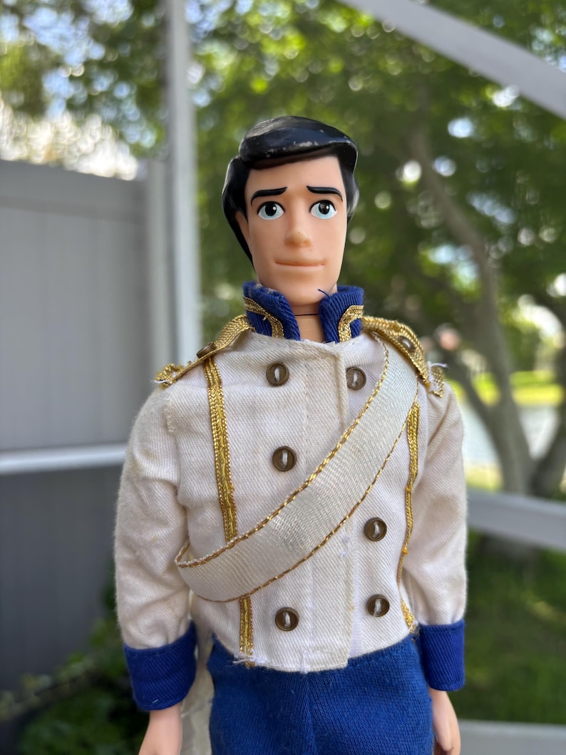 May include: A vintage doll of a prince, wearing a white jacket with gold trim and dark brown buttons. The jacket has a white sash and blue accents. The doll has black hair and is looking forward.