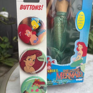 Pode incluir: Um cart&atilde;o vertical com tr&ecirc;s bot&otilde;es circulares com gr&aacute;ficos de A Pequena Sereia, posicionado ao lado de uma boneca Ariel na caixa. Os bot&otilde;es exibem Ariel e Linguado, um close de Ariel e Ariel com o Pr&iacute;ncipe Eric. A caixa da boneca inclui o texto The Little Mermaid e Tyco.