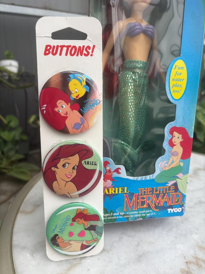 Pode incluir: Um cart&atilde;o vertical com tr&ecirc;s bot&otilde;es circulares com gr&aacute;ficos de A Pequena Sereia, posicionado ao lado de uma boneca Ariel na caixa. Os bot&otilde;es exibem Ariel e Linguado, um close de Ariel e Ariel com o Pr&iacute;ncipe Eric. A caixa da boneca inclui o texto The Little Mermaid e Tyco.