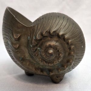 May include: A bronze-toned metal vessel shaped like a nautilus shell. The object features a spiraling, textured exterior with raised ridges and a hollow opening at the top. It stands on three small, rounded feet integrated into the base design.