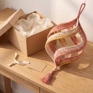 May include: A spiral-shaped crochet ornament featuring alternating bands of dusty rose, cream, and mustard yellow yarn. The decorative piece includes a braided hanging loop at the top, a wooden bead accent, and a matching dusty rose tassel at the bottom.