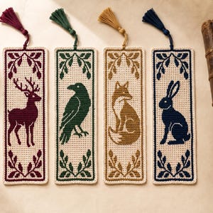 May include: Four rectangular crochet bookmarks in cream yarn, each featuring a different animal silhouette in a distinct color: a maroon deer, a dark green raven, a gold fox, and a navy blue rabbit, all finished with matching tassels.