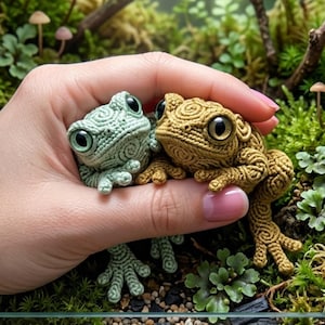 以下が含まれることがあります： 手の中に、緑色と茶色の2匹の精巧なカエルの置物があります。カエルは大きな目と質感のある体を持っています。背景にはミニチュアのキノコと緑があり、ミニチュアガーデンのシーンを作り出しています。