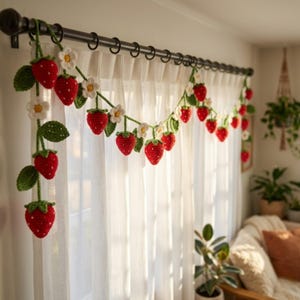 May include: A crochet strawberry garland with red strawberries, green leaves, and white flowers hangs in front of a white curtain. The garland is suspended from a black curtain rod, adding a touch of handmade charm to the room.