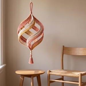 May include: A hanging spiral ornament crafted from crocheted yarn in shades of dusty rose, mustard yellow, and cream. The structure features a twisted, open-work design with a braided loop at the top and a decorative wooden bead and tassel at the bottom.