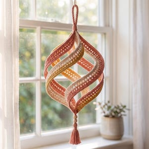 May include: A hanging crochet ornament featuring a spiraling, spherical shape constructed from interconnected bands in shades of dusty pink, cream, and muted yellow. The piece includes a top loop for suspension and a bottom wooden bead with a tassel.