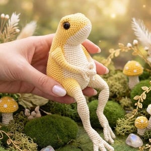 May include: A handmade crochet frog in yellow and cream tones, held in a hand. The frog has large black eyes and is posed with its legs extended. Miniature crochet mushrooms with yellow caps and white spots are in the background.