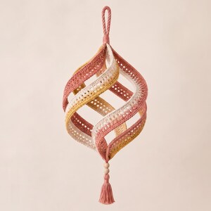 May include: A hanging spiral ornament crafted from crocheted fabric in shades of dusty rose, mustard yellow, and cream. The structure features curved, open panels connected at the top with a loop and at the bottom with a small wooden bead and tassel.