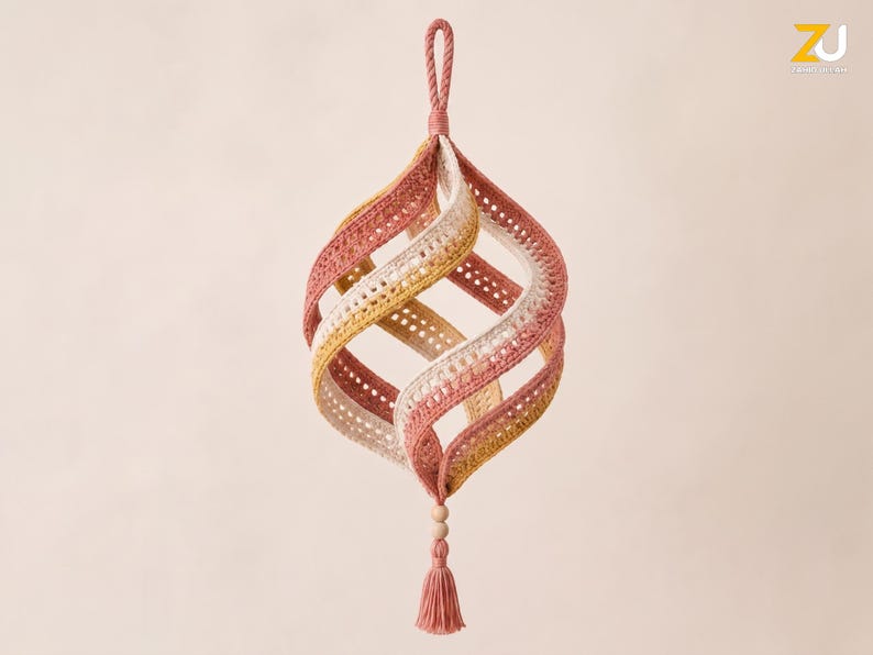 May include: A hanging spiral ornament crafted from crocheted fabric in shades of dusty rose, mustard yellow, and cream. The structure features curved, open panels connected at the top with a loop and at the bottom with a small wooden bead and tassel.