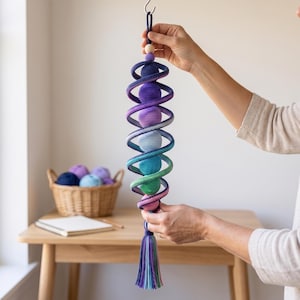 May include: A vertical hanging mobile featuring a spiral structure made of crocheted yarn in a gradient of purple, blue, green, and pink. The spiral surrounds a series of spherical crocheted balls in matching colors, finished with a tassel at the bottom.