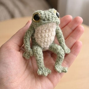 Puede incluir: Una figura de rana de ganchillo hecha a mano en tonos verdes y crema. La rana tiene ojos grandes con centros negros y bordes dorados. La rana está sentada erguida, con los brazos y las piernas extendidos. Es un artículo artesanal caprichoso.