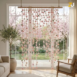 May include: A hanging room divider featuring multiple vertical strands of crocheted pink and white cherry blossom flowers and small green leaves, suspended from a horizontal wooden rod against a large window with a garden view.