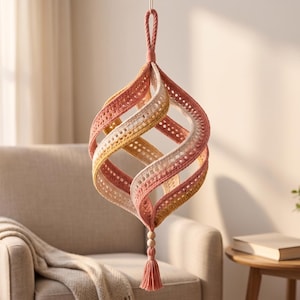 May include: A hanging spiral ornament crafted from crocheted fabric in a gradient of dusty rose, soft yellow, and cream. The structure features a twisted, open-work design with a braided top loop and a bottom tassel accented by two wooden beads.