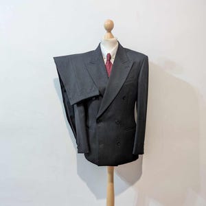 Puede incluir: Un traje gris oscuro de doble botonadura exhibido en un maniqu&iacute; de madera. El traje incluye una chaqueta y un pantal&oacute;n a juego, combinados con una camisa blanca y una corbata roja estampada. El maniqu&iacute; est&aacute; sobre un soporte de madera clara.