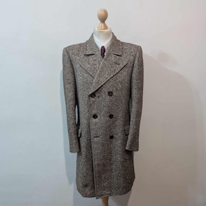 Puede incluir: Abrigo de tweed marrón y gris de doble botonadura, con botones oscuros y solapa con muescas. El abrigo se exhibe en un maniquí de madera, revelando una camisa blanca con cuello y una corbata estampada. Diseño clásico y a medida.