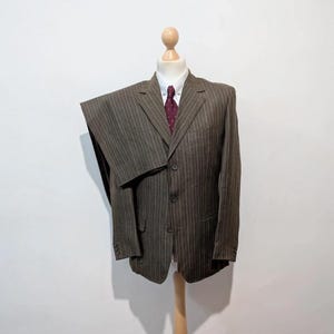 May include: A brown pinstripe suit displayed on a wooden mannequin. The suit includes a jacket, pants, a white dress shirt, and a burgundy tie. The suit is made of a textured fabric and has a classic, tailored design.