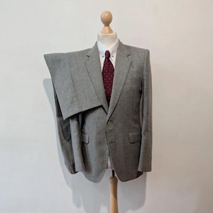 May include: A gray pinstripe suit displayed on a wooden mannequin. The suit includes a blazer with a single button closure and a pair of trousers. A white collared shirt and a burgundy tie with white polka dots complete the ensemble. The mannequin is on a wooden stand.