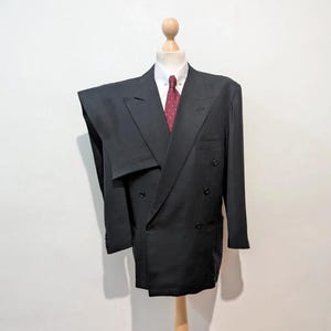 May include: A black double-breasted suit displayed on a wooden mannequin. The suit includes a white collared shirt and a red patterned tie. The mannequin is on a wooden stand, set against a white wall and a terracotta tile floor.
