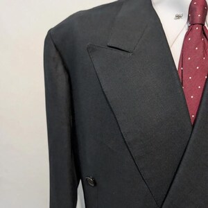 May include: A black double-breasted suit jacket with a white collared shirt and a burgundy tie with white polka dots. A silver lapel pin is visible on the shirt. The suit jacket has two rows of buttons.