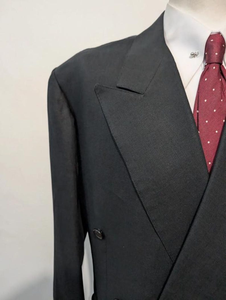 May include: A black double-breasted suit jacket with a white collared shirt and a burgundy tie with white polka dots. A silver lapel pin is visible on the shirt. The suit jacket has two rows of buttons.