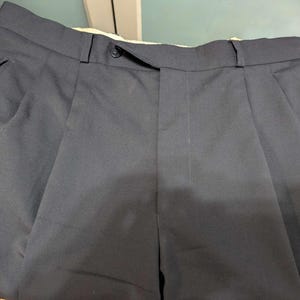 May include: Close-up of navy blue pleated trousers. The trousers feature a button closure and side pockets. The fabric appears to be a smooth, woven material. The trousers are neatly folded, showcasing the tailored design.