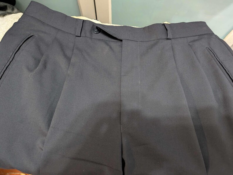 May include: Close-up of navy blue pleated trousers. The trousers feature a button closure and side pockets. The fabric appears to be a smooth, woven material. The trousers are neatly folded, showcasing the tailored design.