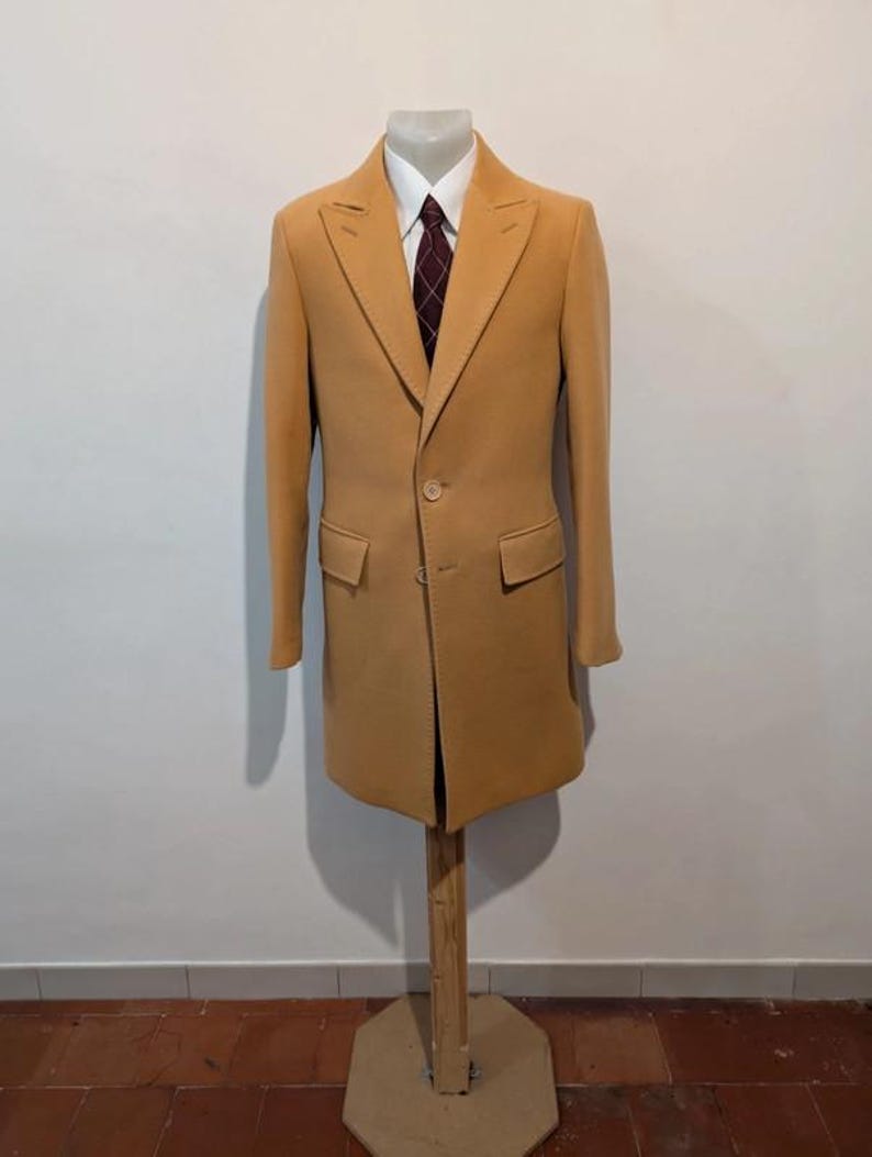 Puede incluir: Un abrigo color canela exhibido en un maniqu&iacute;. El abrigo tiene un dise&ntilde;o cl&aacute;sico con solapa de muesca, dos bolsillos delanteros y cierre de un solo bot&oacute;n. Debajo del abrigo se ven una camisa blanca y una corbata burdeos estampada.