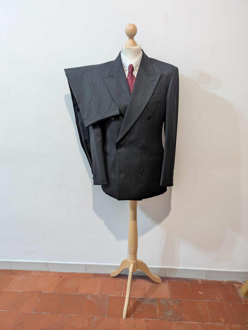 Puede incluir: Un traje gris oscuro de doble botonadura exhibido en un maniqu&iacute; de madera. El traje incluye una chaqueta y un pantal&oacute;n a juego, combinados con una camisa blanca y una corbata roja estampada. El maniqu&iacute; est&aacute; sobre un soporte de madera clara.