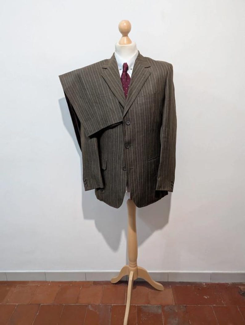 May include: A brown pinstripe suit displayed on a wooden mannequin. The suit includes a jacket, pants, a white dress shirt, and a burgundy tie. The suit is made of a textured fabric and has a classic, tailored design.