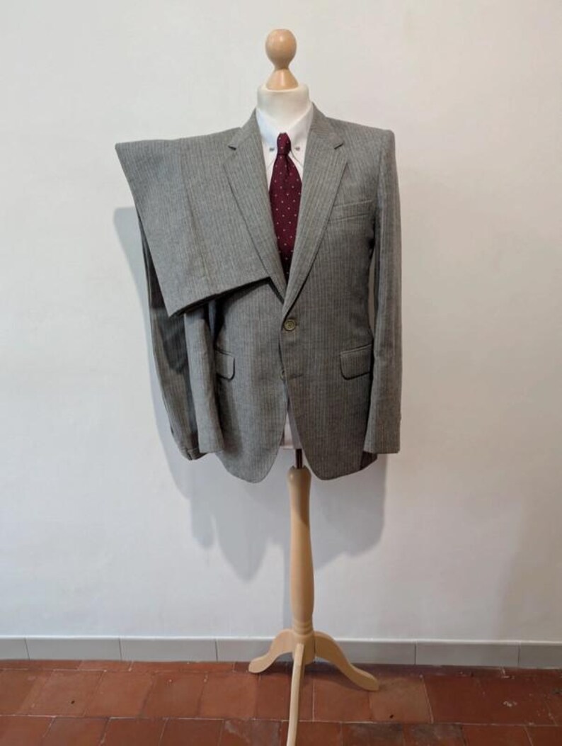May include: A gray pinstripe suit displayed on a wooden mannequin. The suit includes a blazer with a single button closure and a pair of trousers. A white collared shirt and a burgundy tie with white polka dots complete the ensemble. The mannequin is on a wooden stand.