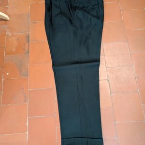 May include: Black dress pants with a pleated front and cuffed hem. The pants are made of a solid, dark fabric and are laid flat on a tiled surface. The pants have a button and zipper closure.