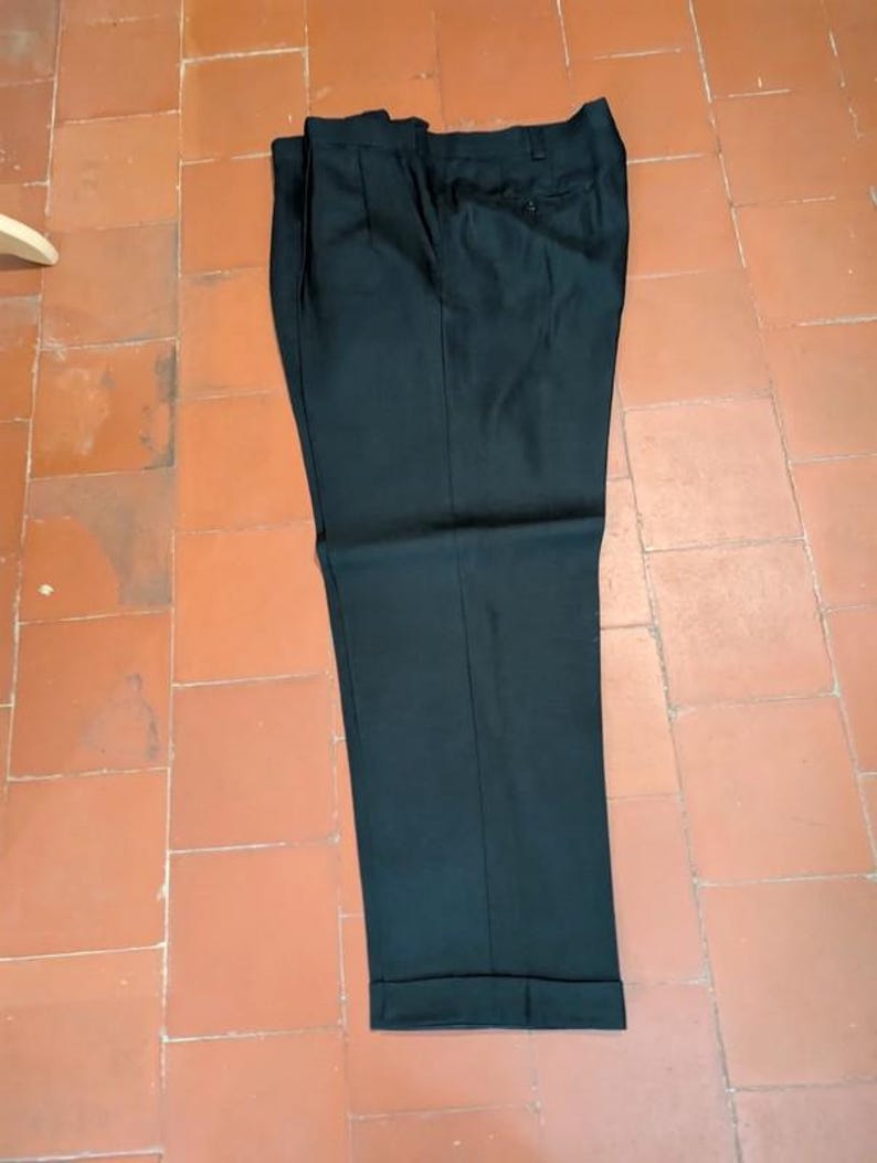 May include: Black dress pants with a pleated front and cuffed hem. The pants are made of a solid, dark fabric and are laid flat on a tiled surface. The pants have a button and zipper closure.