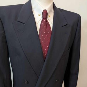 May include: A navy blue double-breasted suit jacket, white shirt, and burgundy tie with white spots. The jacket has a notched lapel and two buttons. The tie is secured with a gold tie pin.