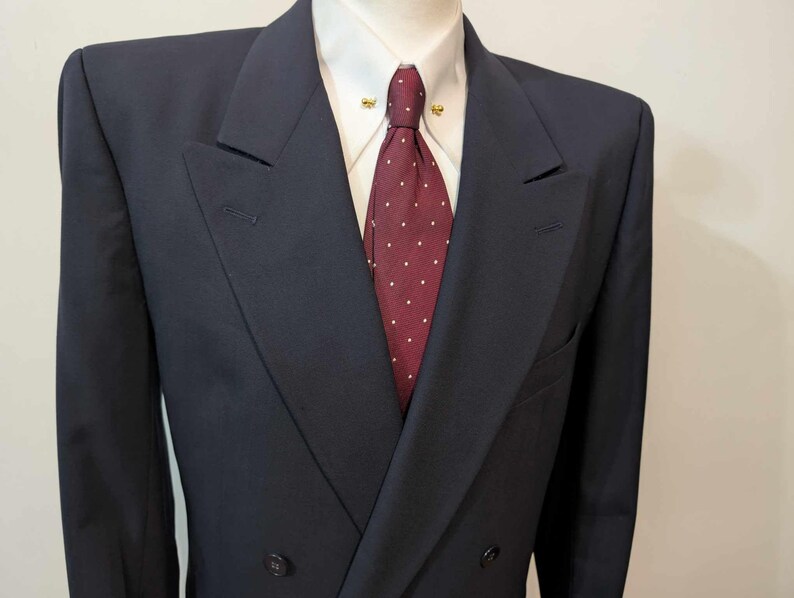 May include: A navy blue double-breasted suit jacket, white shirt, and burgundy tie with white spots. The jacket has a notched lapel and two buttons. The tie is secured with a gold tie pin.