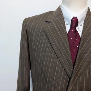May include: A brown pinstripe suit jacket with a white dress shirt and a burgundy tie with white polka dots. The suit is displayed on a mannequin. The tie has a tie pin.