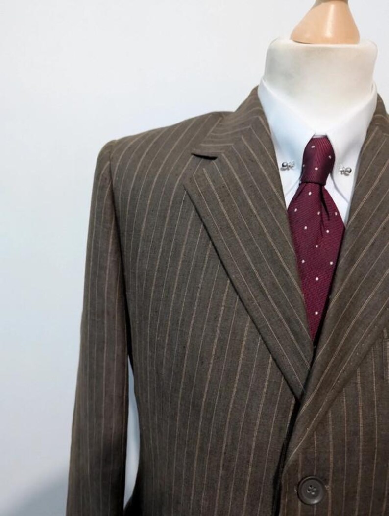 May include: A brown pinstripe suit jacket with a white dress shirt and a burgundy tie with white polka dots. The suit is displayed on a mannequin. The tie has a tie pin.
