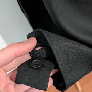 May include: Close-up of a black garment cuff with a button and buttonhole. The fabric appears to be a woven material. The button is a dark, round shape.