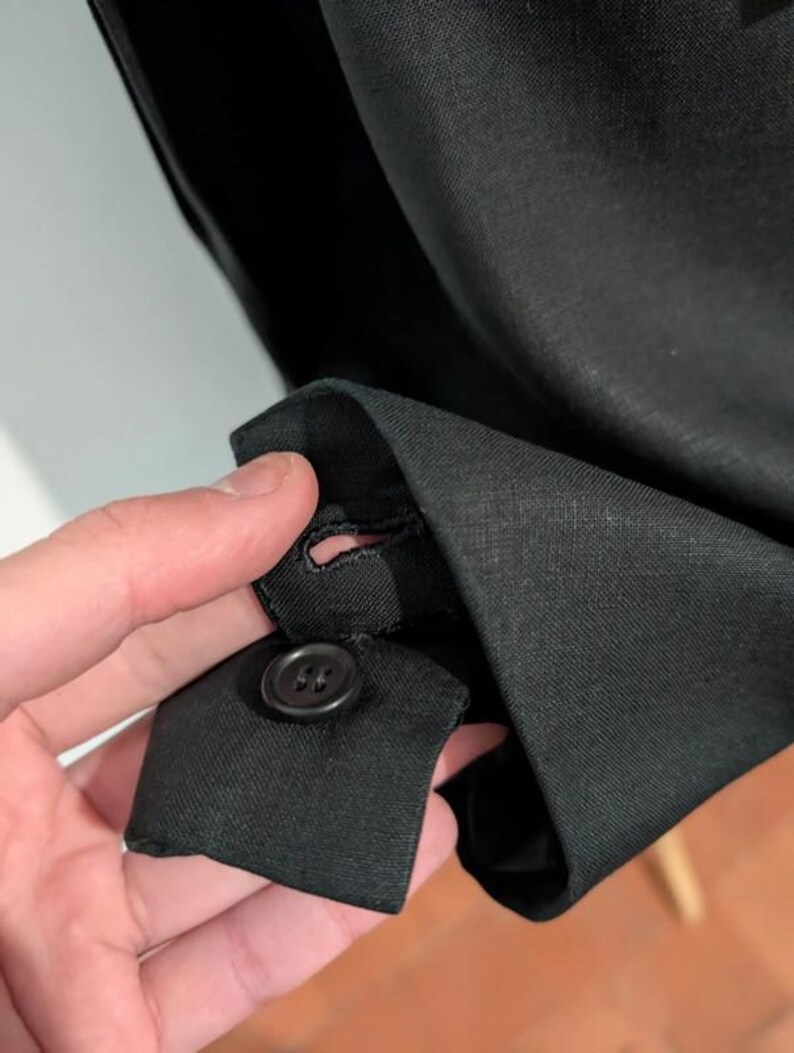 May include: Close-up of a black garment cuff with a button and buttonhole. The fabric appears to be a woven material. The button is a dark, round shape.
