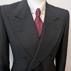 Puede incluir: Un traje cruzado gris oscuro con un sutil patr&oacute;n cuadrado. El traje presenta una camisa blanca con cuello, una corbata burdeos con lunares blancos y alfileres de cuello dorados. La chaqueta tiene dos filas de botones y un dise&ntilde;o de solapa cl&aacute;sico.