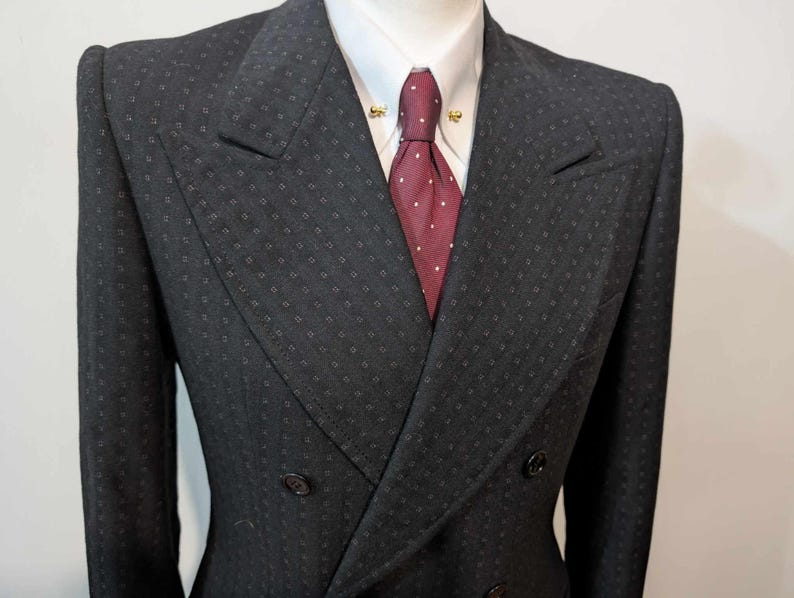 Puede incluir: Un traje cruzado gris oscuro con un sutil patr&oacute;n cuadrado. El traje presenta una camisa blanca con cuello, una corbata burdeos con lunares blancos y alfileres de cuello dorados. La chaqueta tiene dos filas de botones y un dise&ntilde;o de solapa cl&aacute;sico.