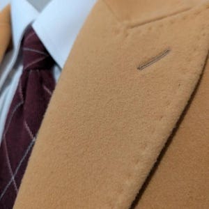 Puede incluir: Primer plano de una blazer color beige con una camisa blanca con cuello y una corbata a cuadros burdeos. La blazer tiene un ojal y una tela texturizada. La corbata presenta un patr&oacute;n de cuadros cl&aacute;sico.