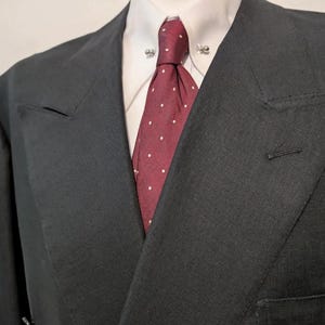 May include: A dark gray double-breasted suit jacket with a white collared shirt and a red tie with white polka dots. The jacket has a notched lapel and a single button closure. The tie is secured with a tie pin.
