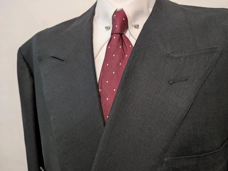 May include: A dark gray double-breasted suit jacket with a white collared shirt and a red tie with white polka dots. The jacket has a notched lapel and a single button closure. The tie is secured with a tie pin.