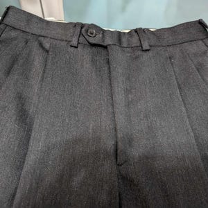Puede incluir: Primer plano de un pantal&oacute;n plisado gris oscuro. El pantal&oacute;n tiene cierre de bot&oacute;n y trabillas para el cintur&oacute;n. La tela tiene una sutil textura vertical. Los pantalones probablemente forman parte de un traje o vestimenta formal.